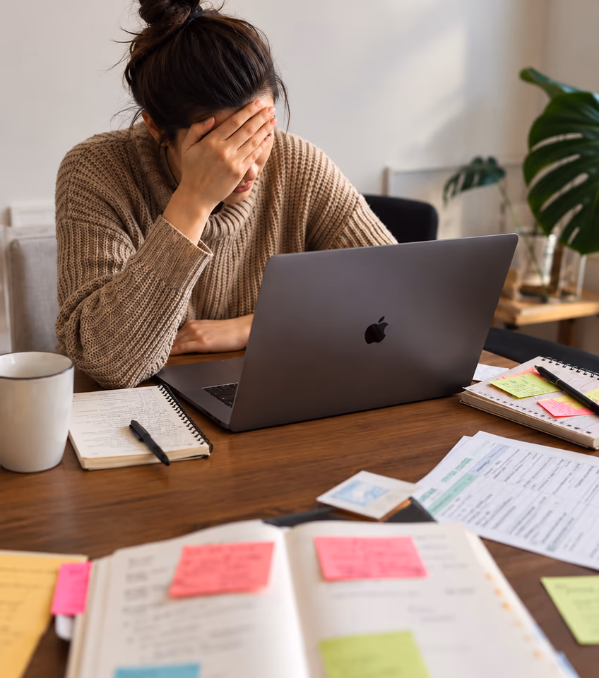 A woman with her face in her hand at a laptop, surrounded by notes